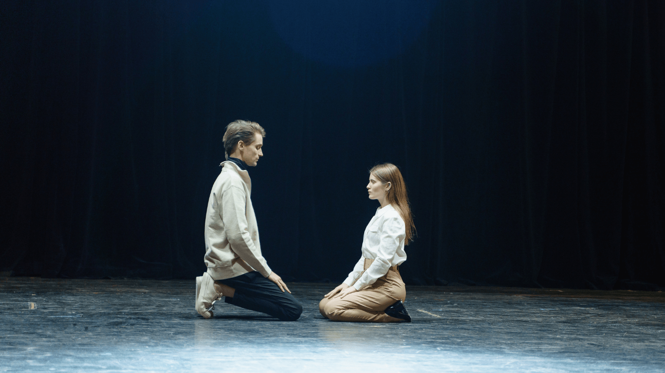 A man and woman sit on the floor, facing a stage, engaged in conversation or enjoying a performance